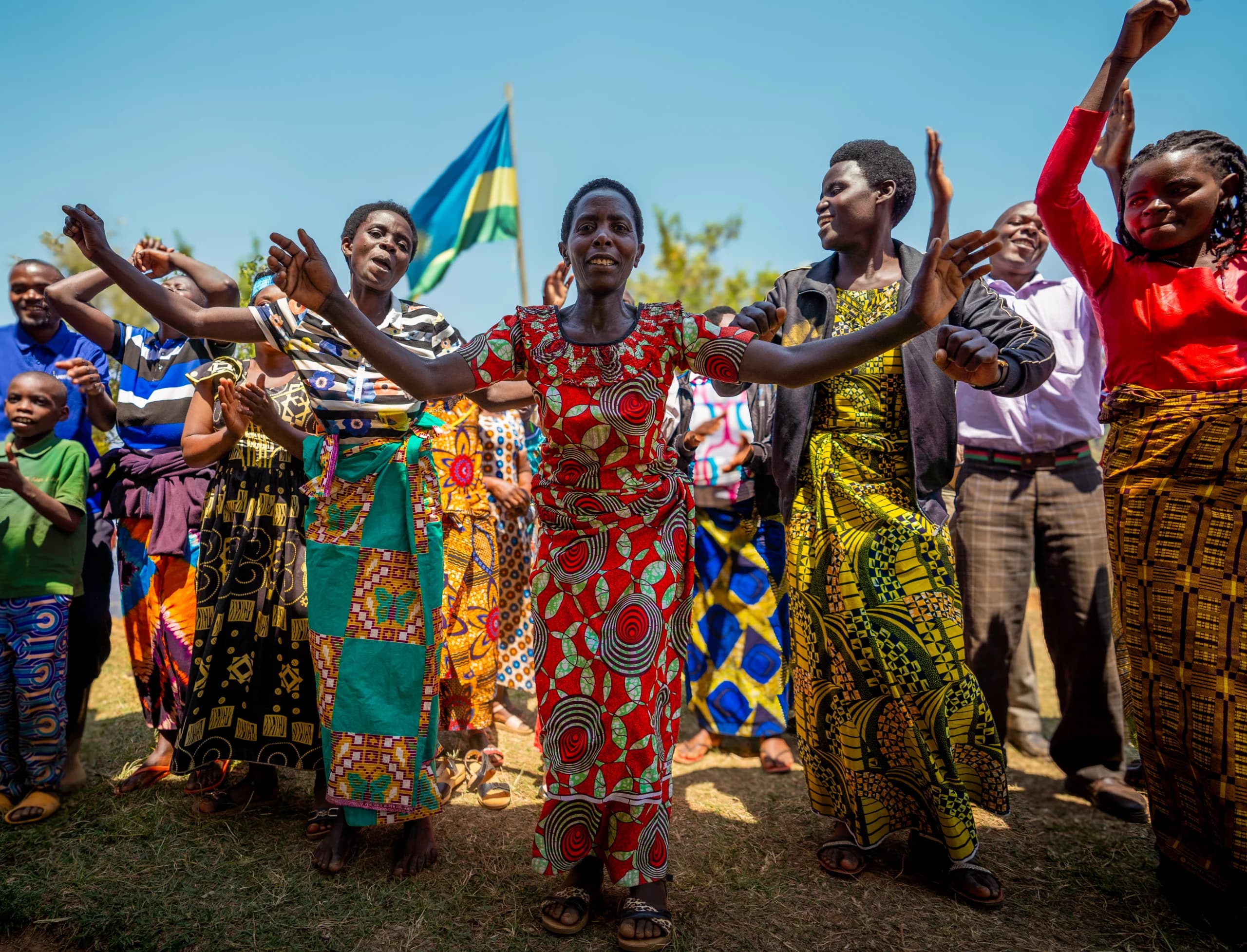 Women in bright printed dresses dance with raised arms on dry grass, Rwandan flag behind against blue sky