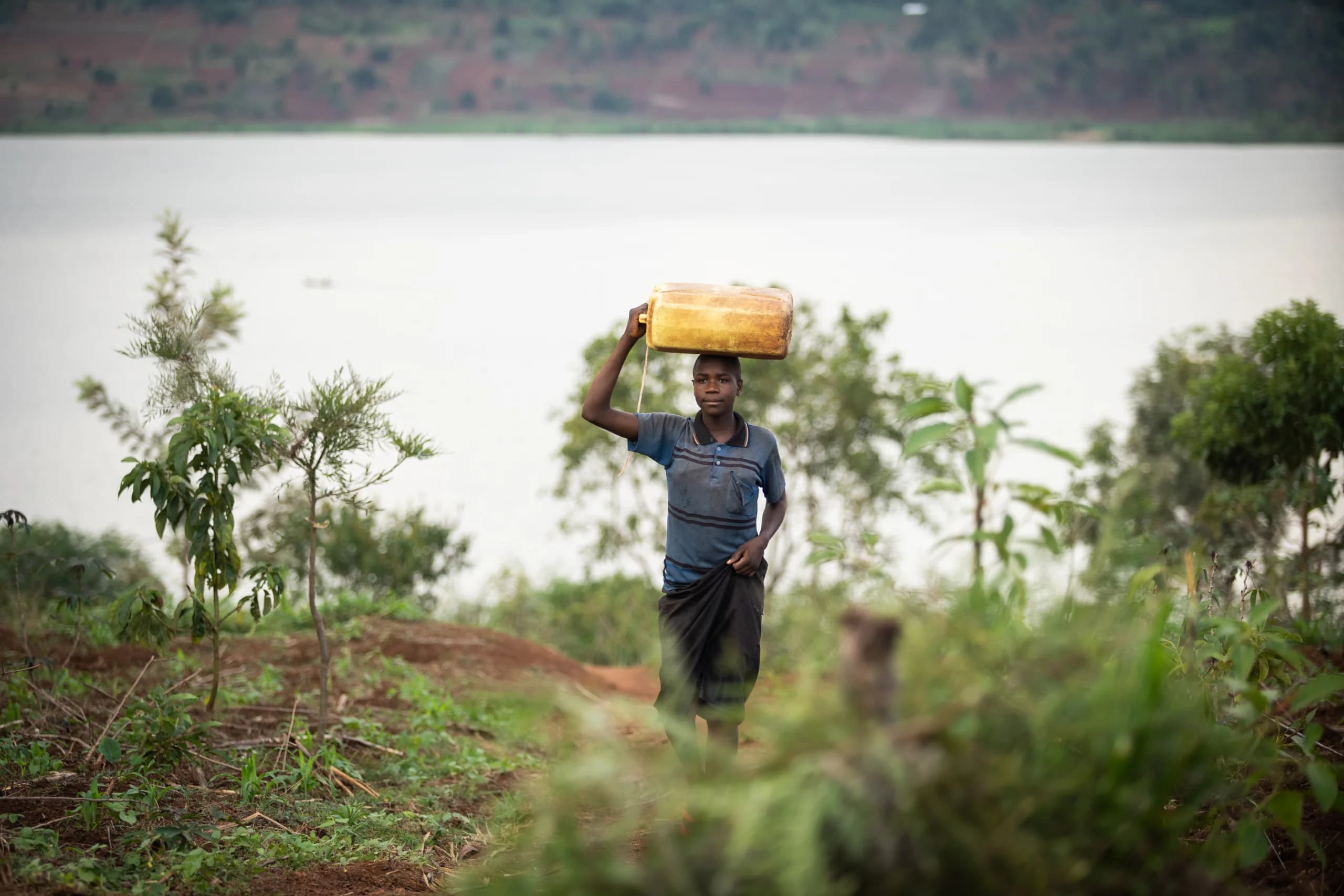 Boy carrying a yellow jerrycan of water on his head along a path above a lake