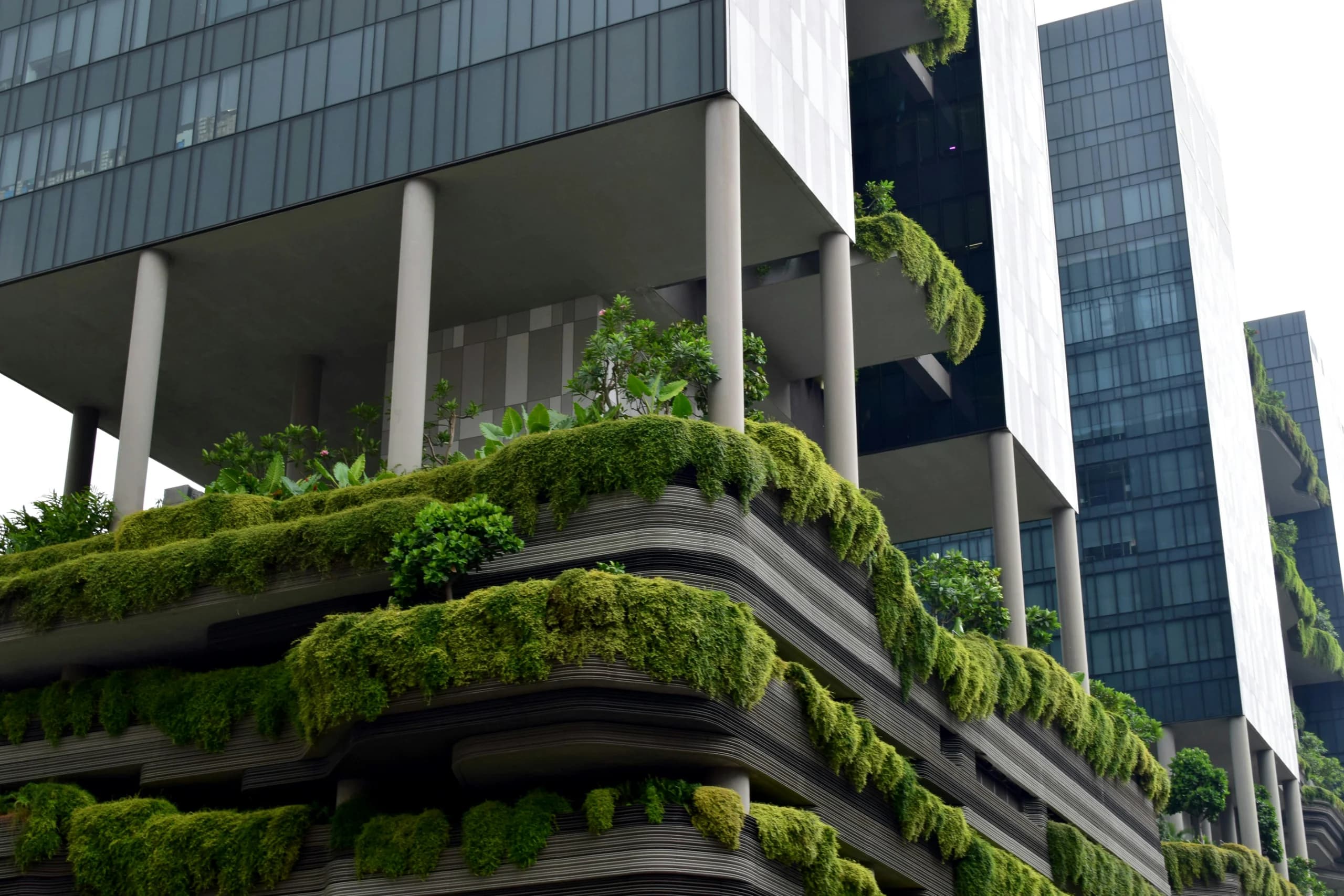 Tiered planted terraces of cascading greenery on a high-rise building, with glass towers behind under overcast sky