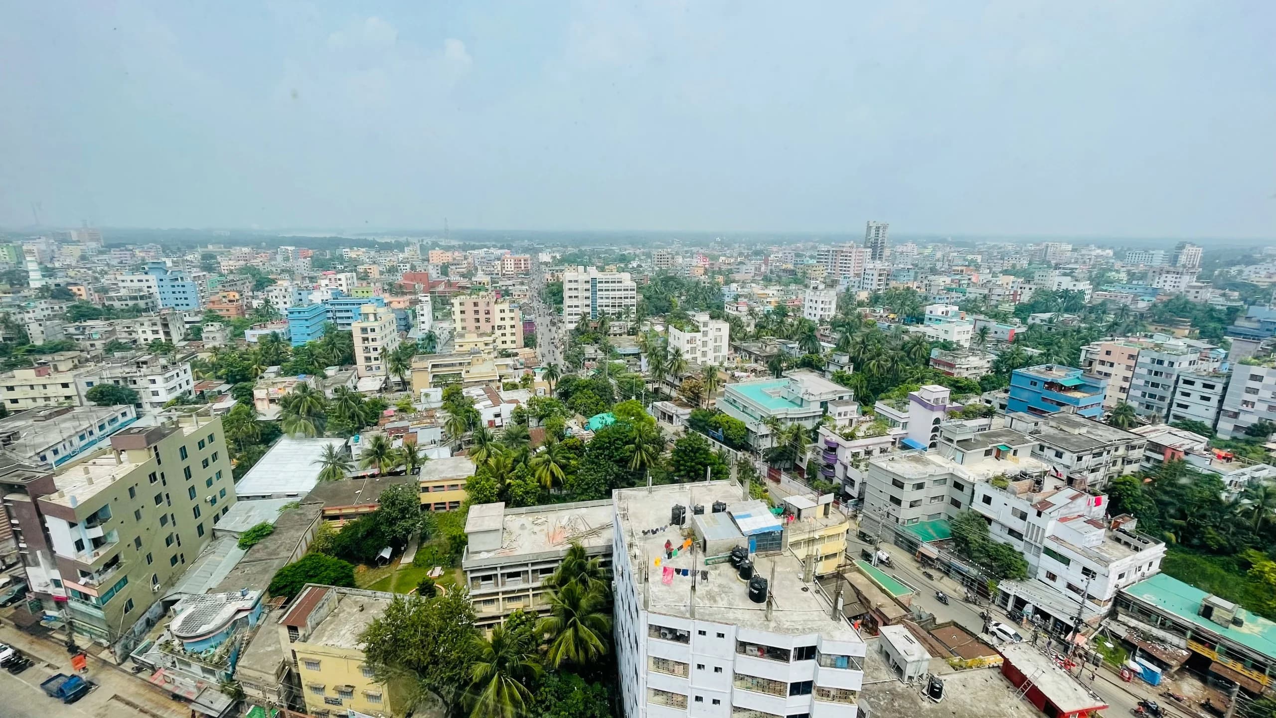High vantage view over a dense South Asian city of low concrete blocks, palm trees and rooftops under hazy sky