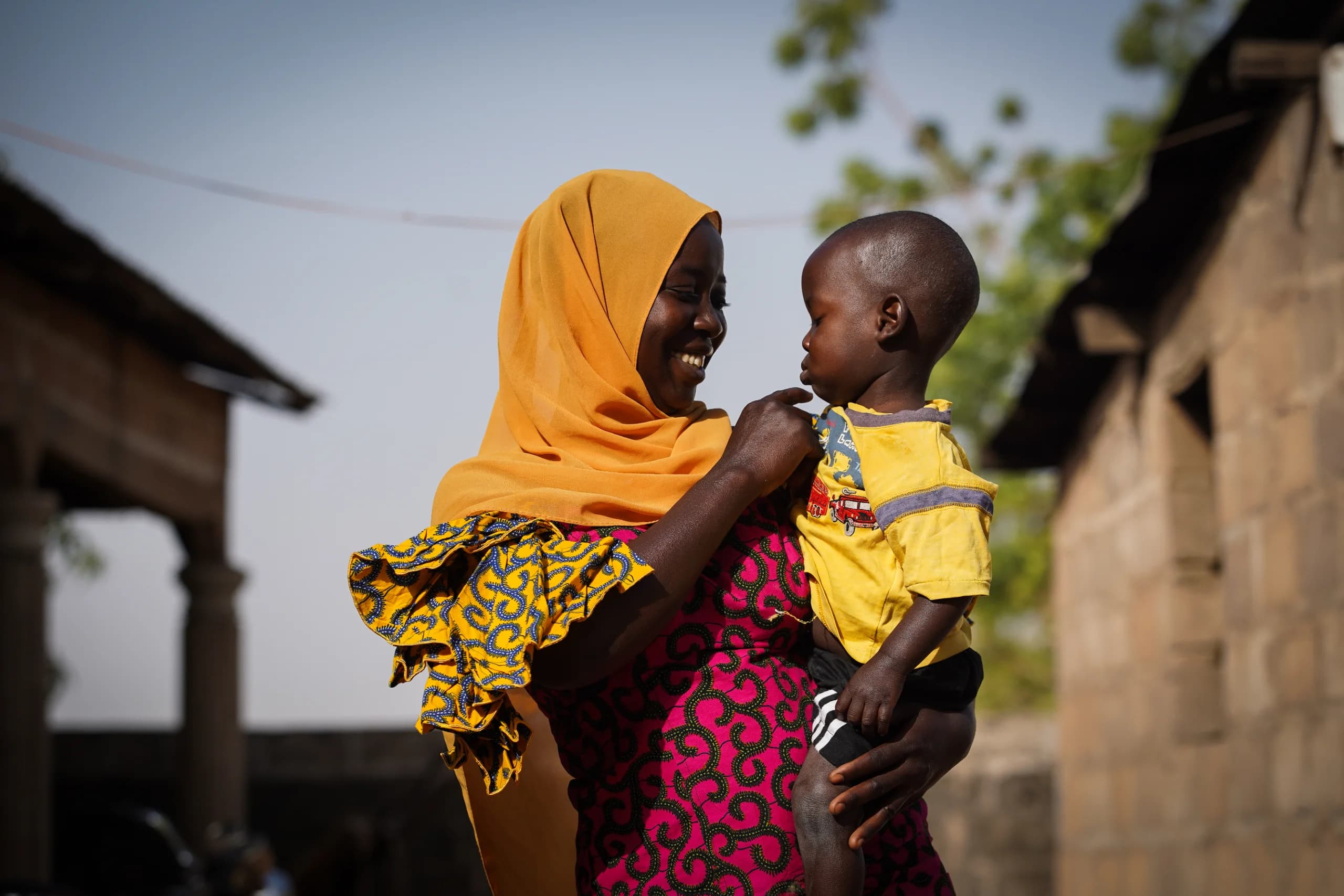 Smiling woman in yellow headscarf and pink patterned dress holds a toddler in a yellow shirt outdoors in a village