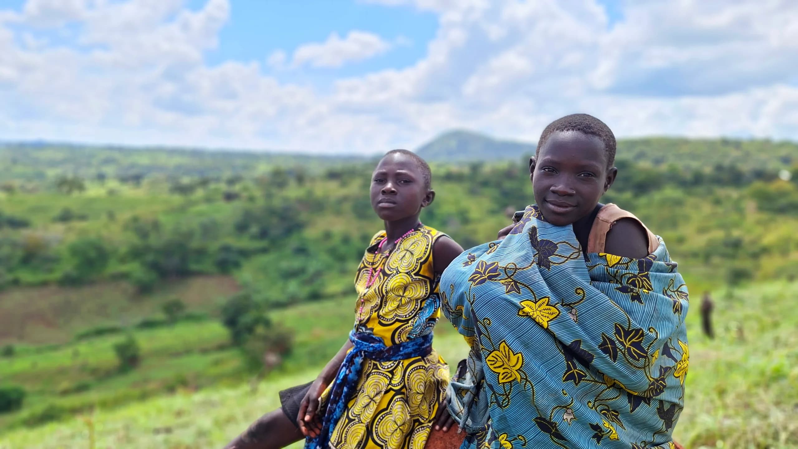 Two girls in patterned wax-print cloth sit on a grassy hillside with green valley and hills behind