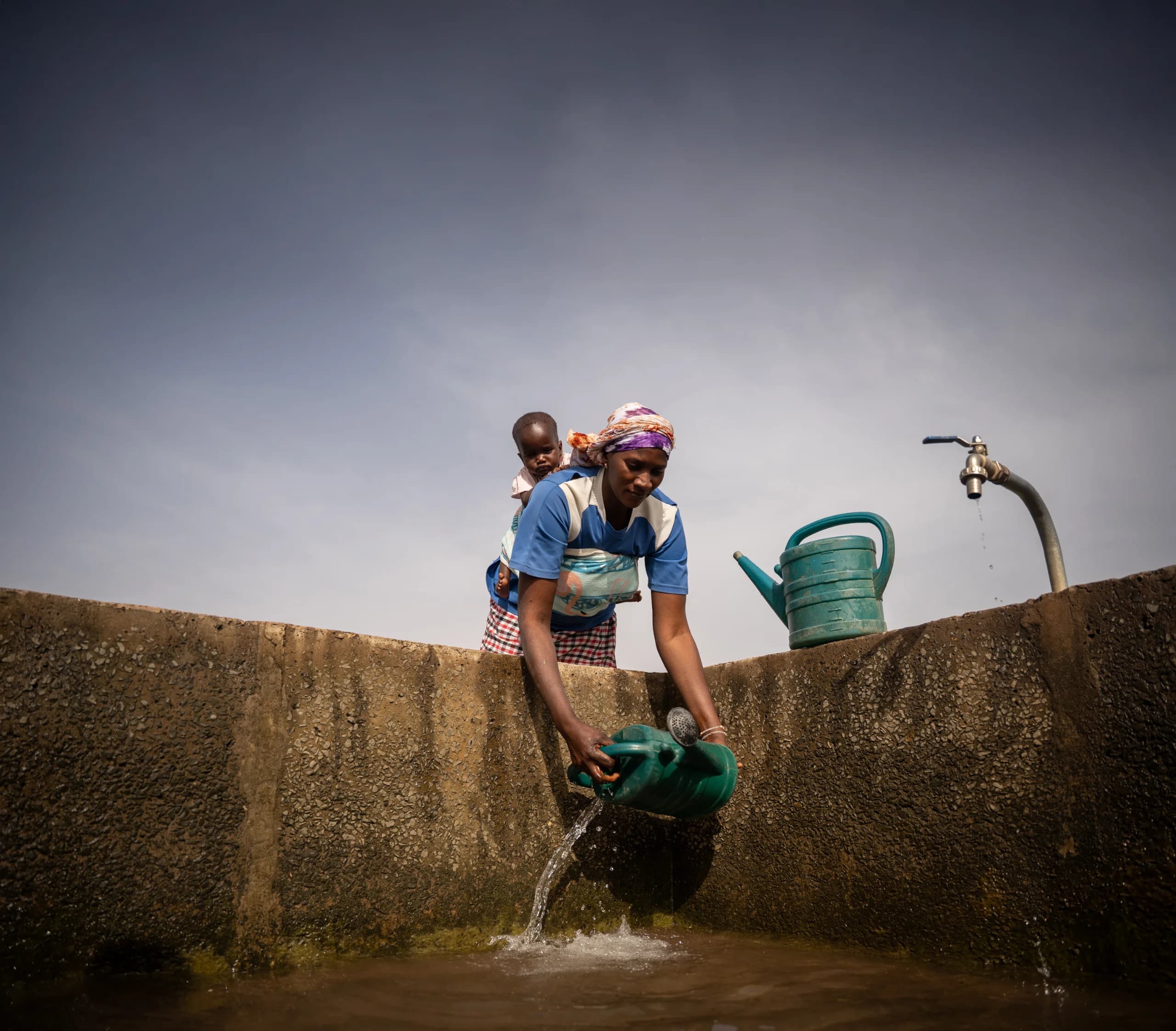 Woman with a baby on her back leans over a concrete tank wall, pouring water from a green watering can