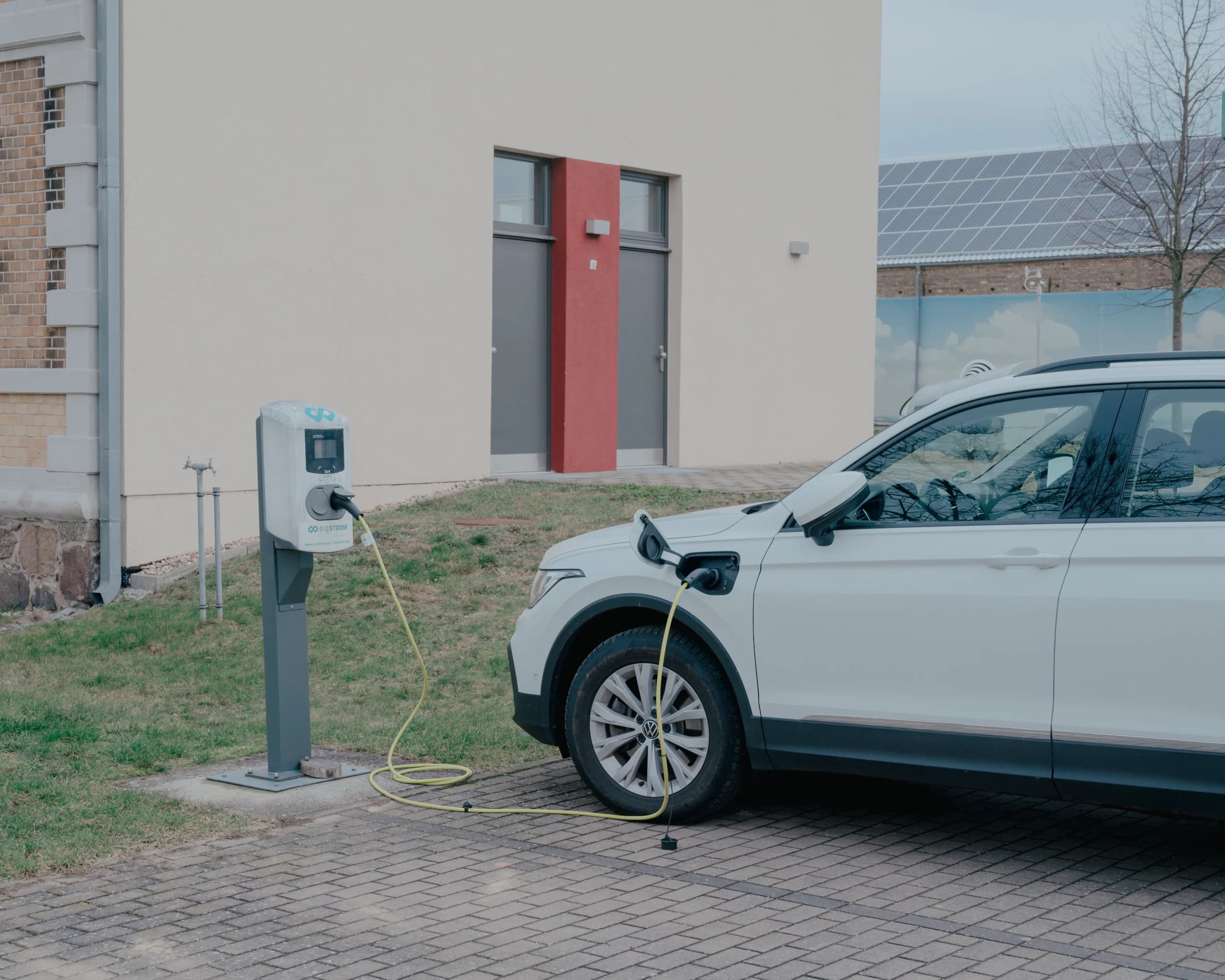 White VW SUV plugged into a kerbside charging post by a building, yellow cable across paving, solar roof behind