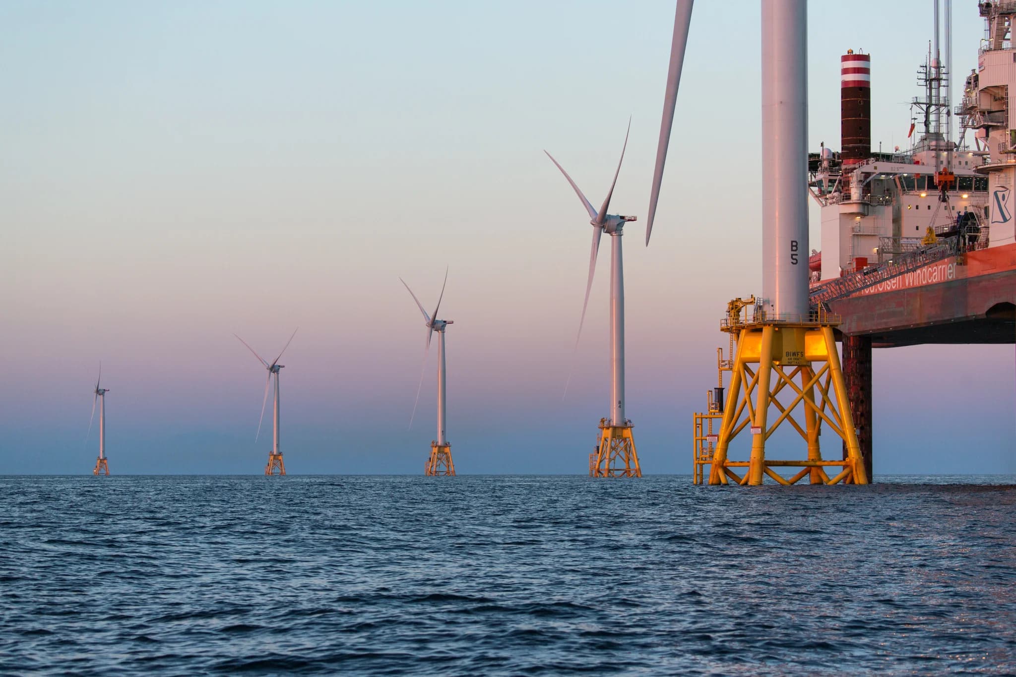 Row of five offshore wind turbines on yellow jacket foundations at dusk, with an installation vessel alongside the nearest tower