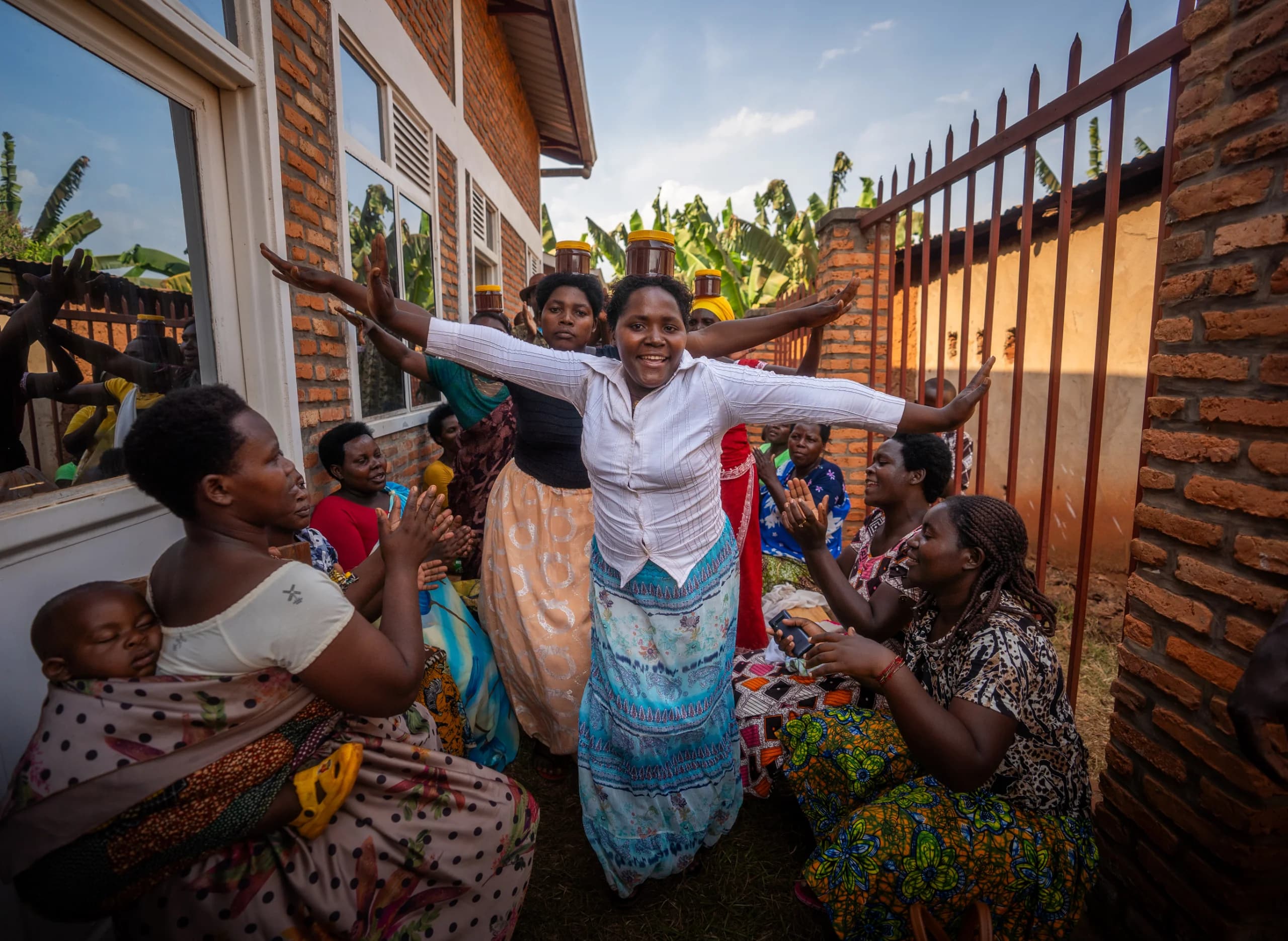 Smiling woman with arms outstretched dances among clapping women in a yard, some balancing honey jars on their heads